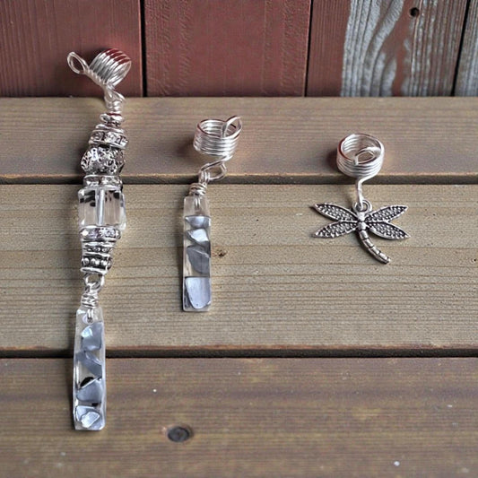 Three silver hair accessories are displayed on a wooden surface, including a clear glass pendant, a silver spiral clip, and a silver dragonfly charm.