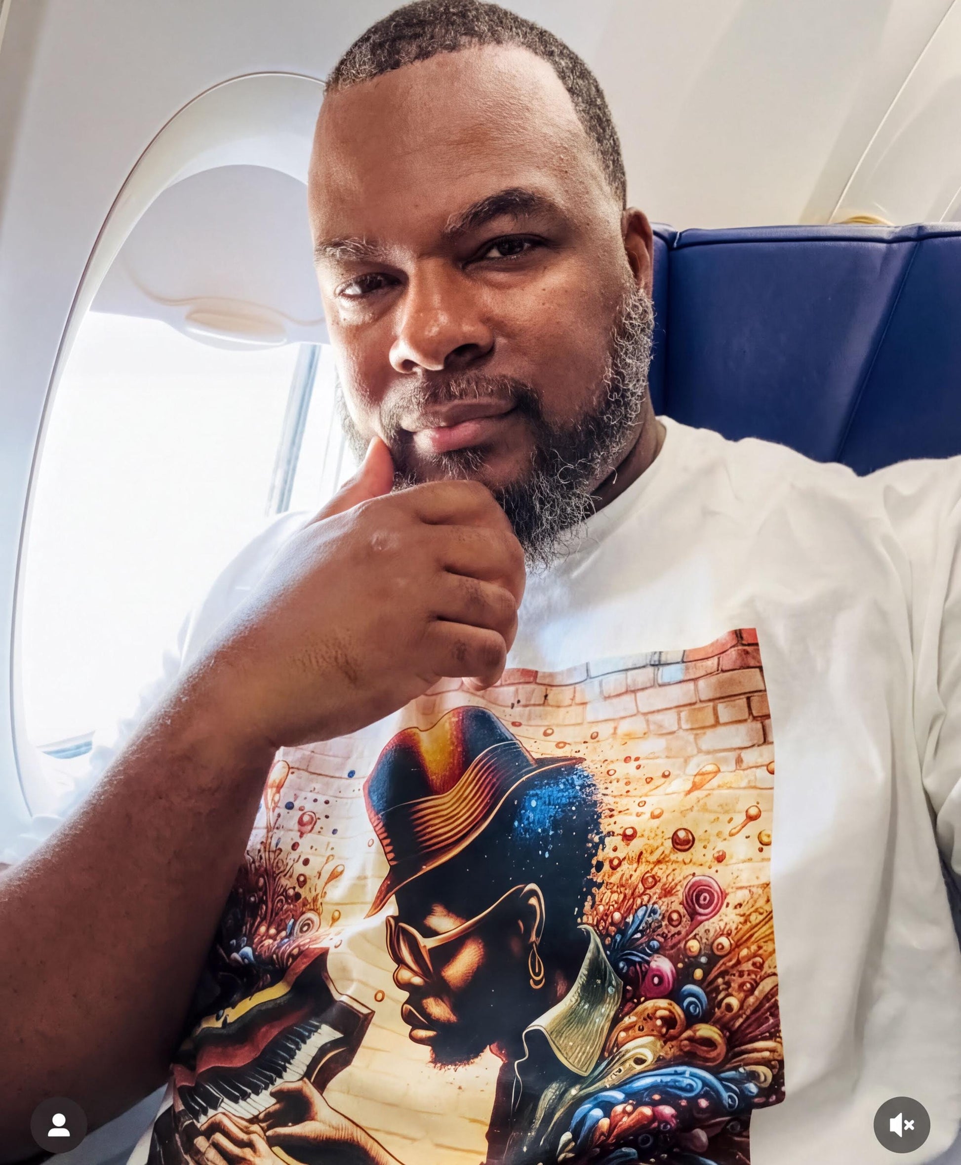 A man wearing a white t-shirt with a colorful graphic design is sitting in an airplane, looking out the window.
