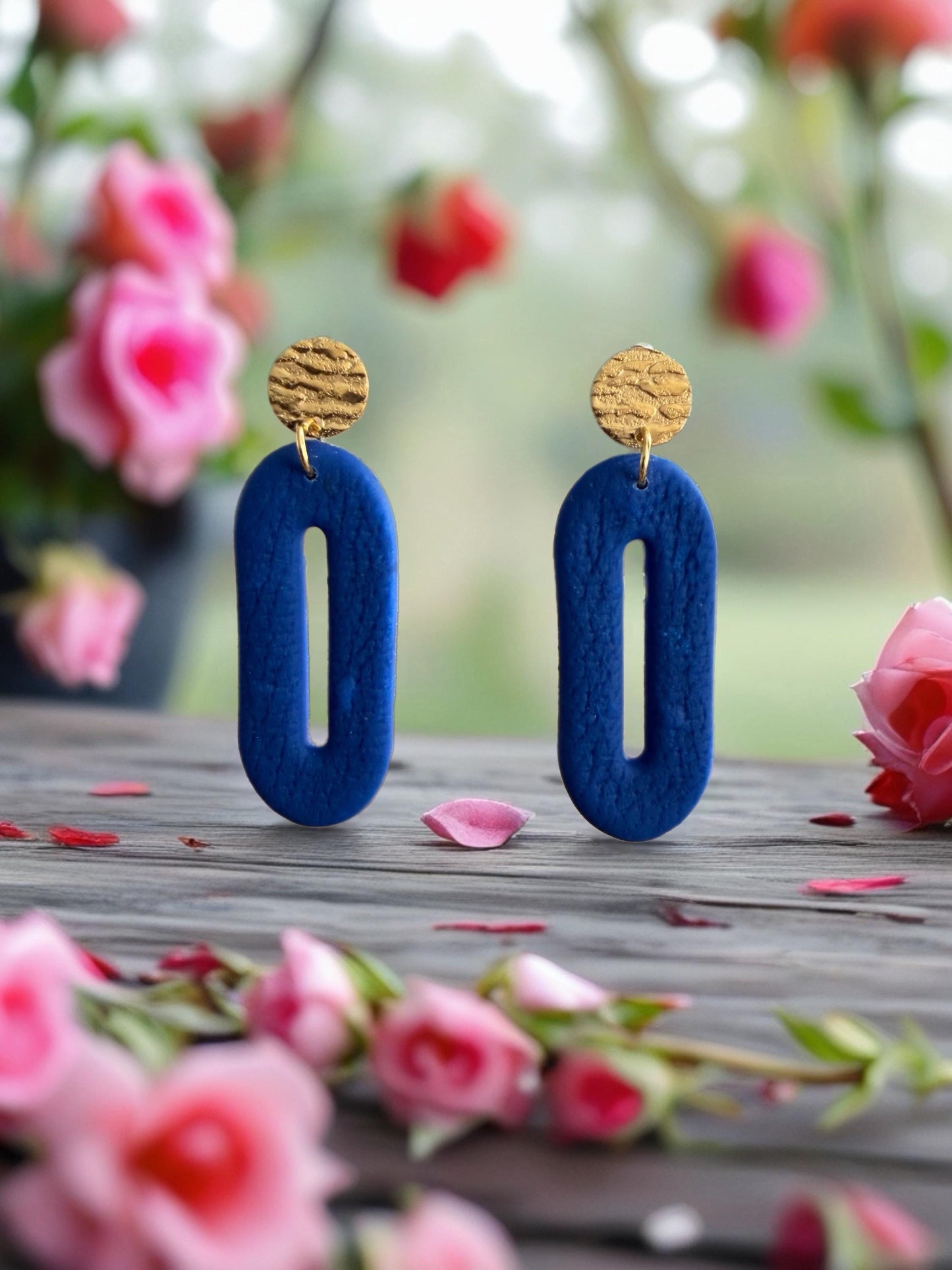 a pair of blue earrings with gold accents, placed on a wooden surface surrounded by pink flowers.