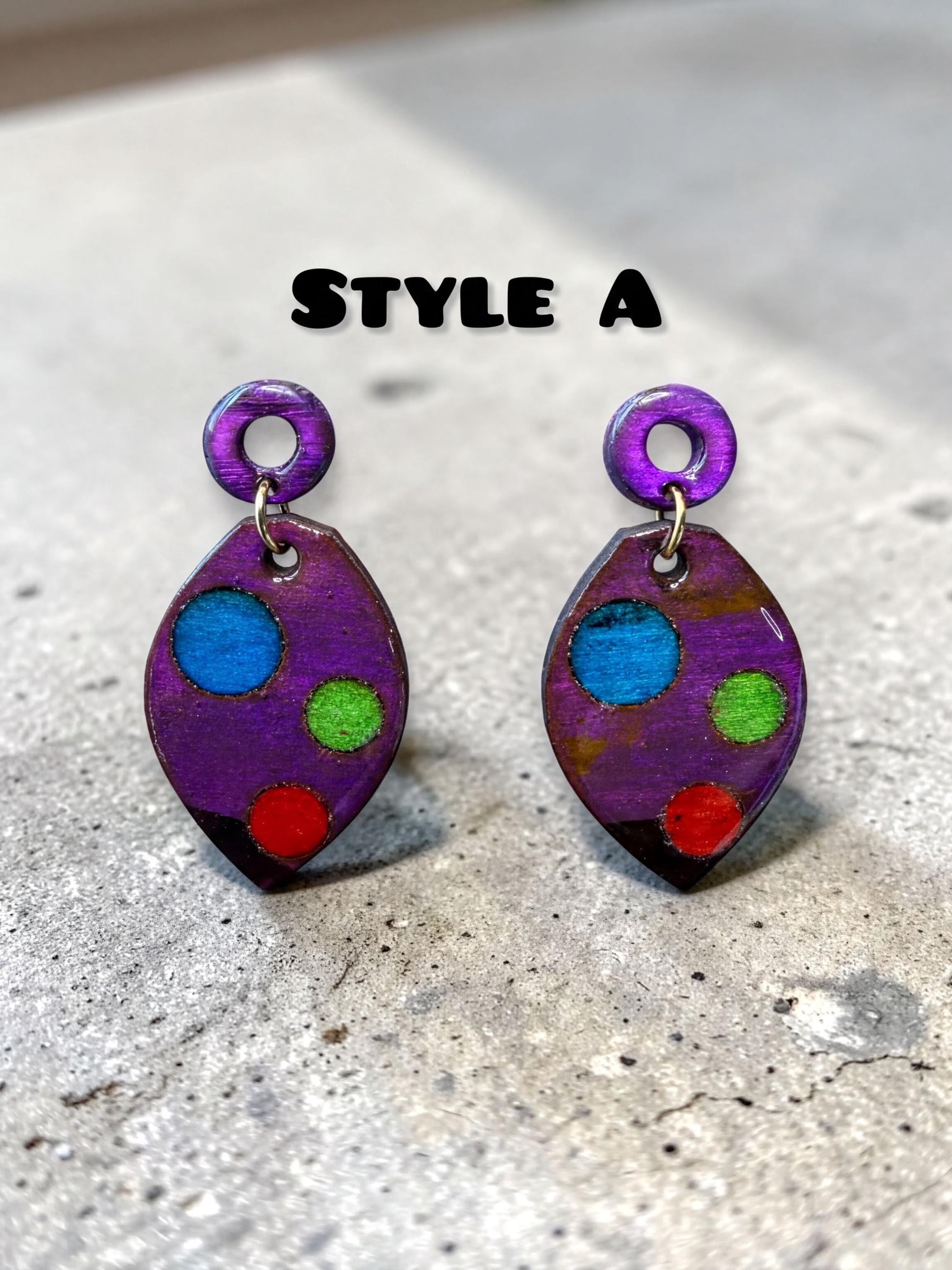 two purple earrings with colorful polka dots, placed on a concrete surface.