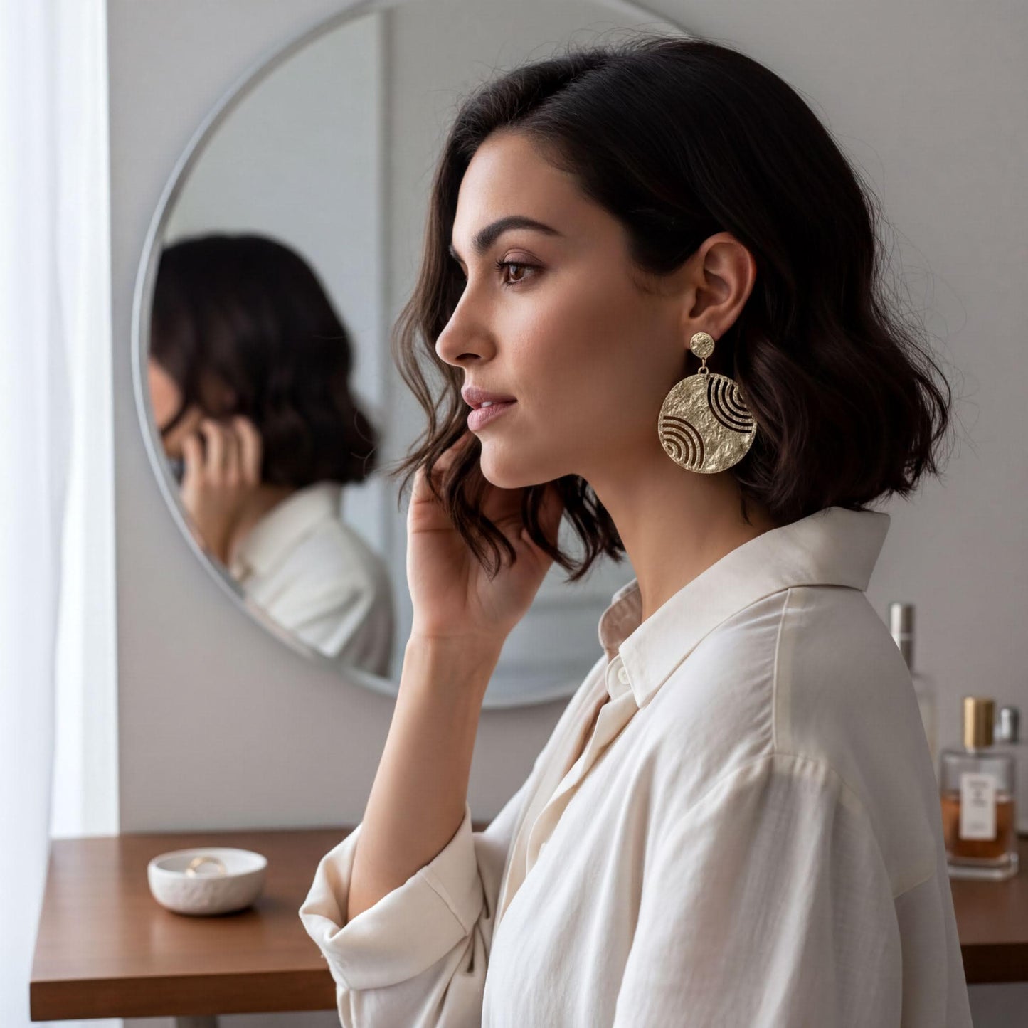 A woman with dark hair wearing a white blouse and large gold earrings is seen in a mirror, with her hand touching her face.