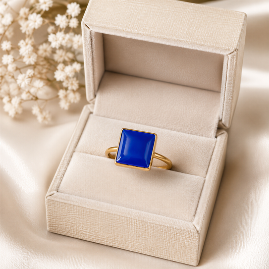 A gold ring with a blue gemstone is displayed in a white jewelry box, placed on a white fabric background.