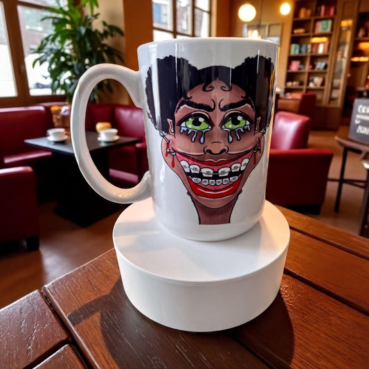 A white mug with a colorful, cartoonish face design featuring a smiling mouth, green eyes, and a large nose is displayed on a wooden table.