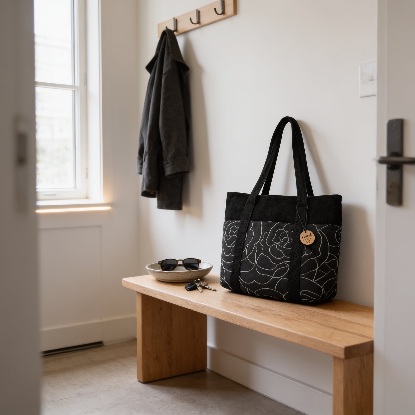A wooden bench with a black and white tote bag and a bowl of sunglasses on it, situated in a hallway with a coat rack and a window.