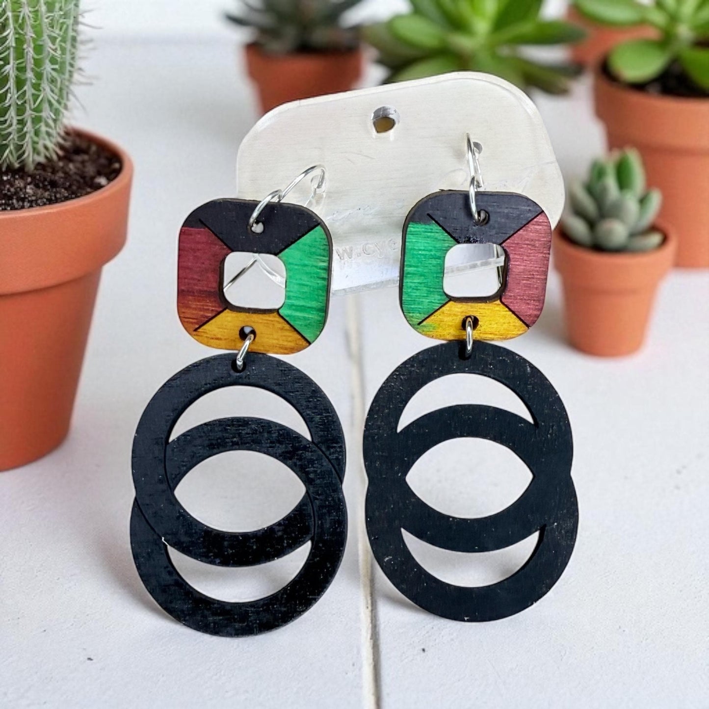 A pair of earrings with black circular cutouts and colorful geometric shapes, displayed on a white surface with potted plants in the background.