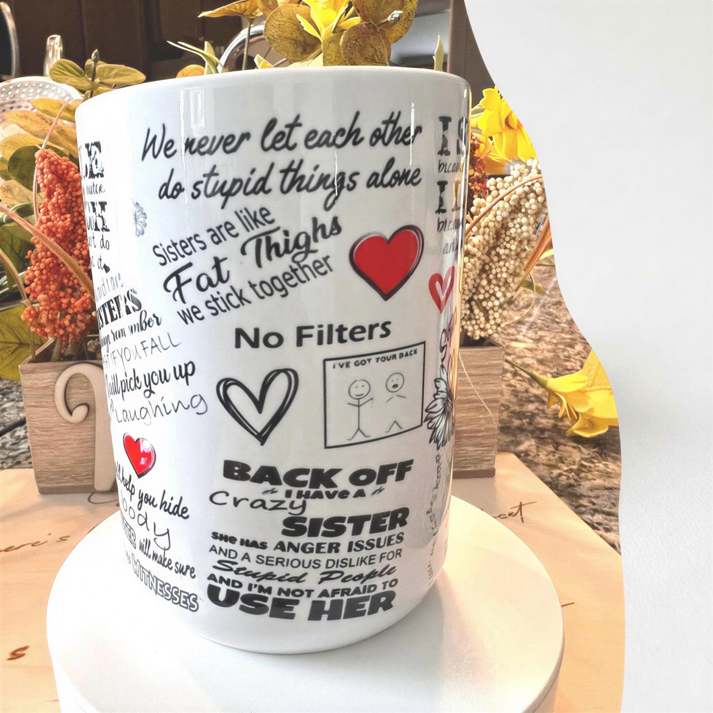 a white mug with various phrases and illustrations printed on it, placed on a table with a vase of flowers in the background.