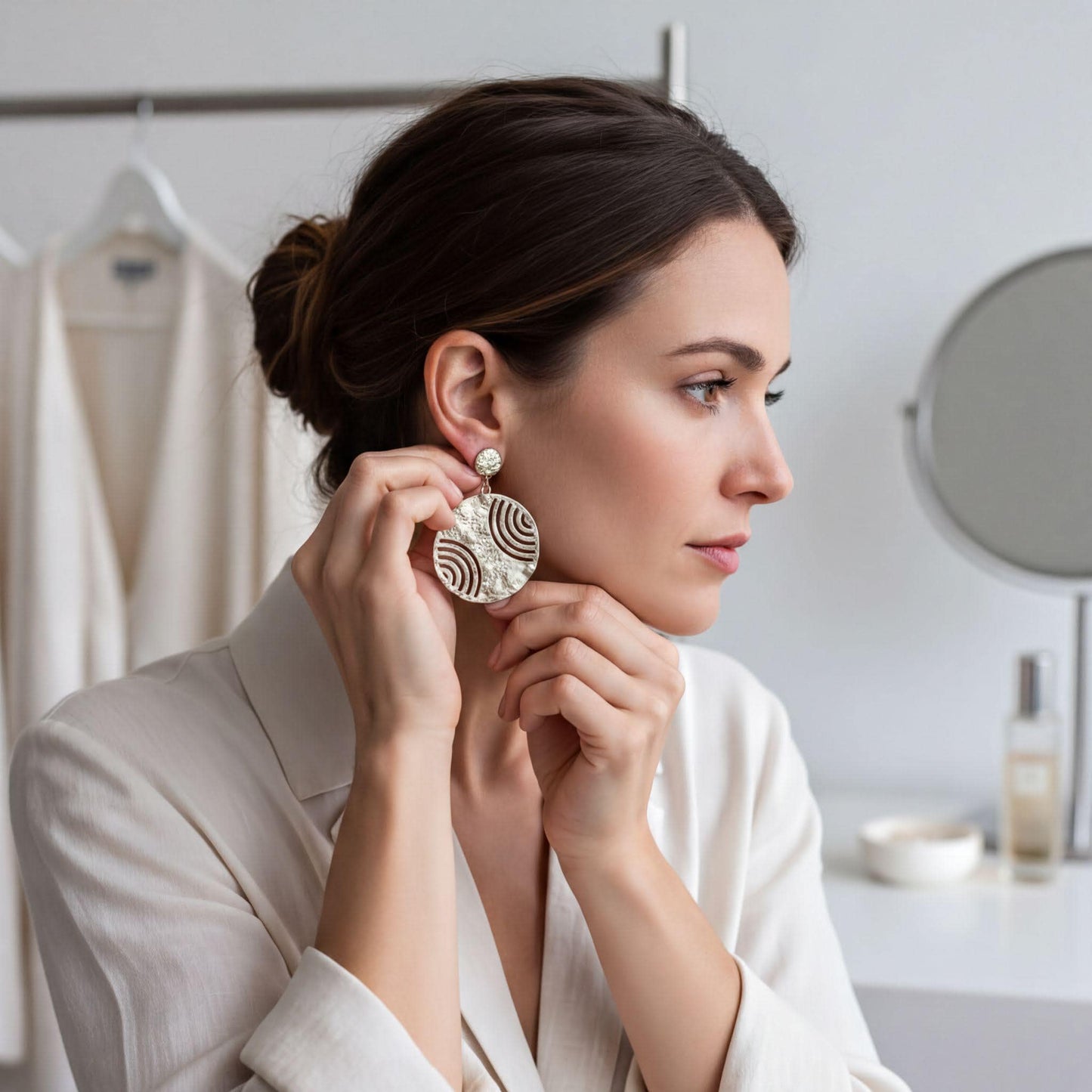 A woman in a white blazer is adjusting her large, ornate earrings in front of a mirror.